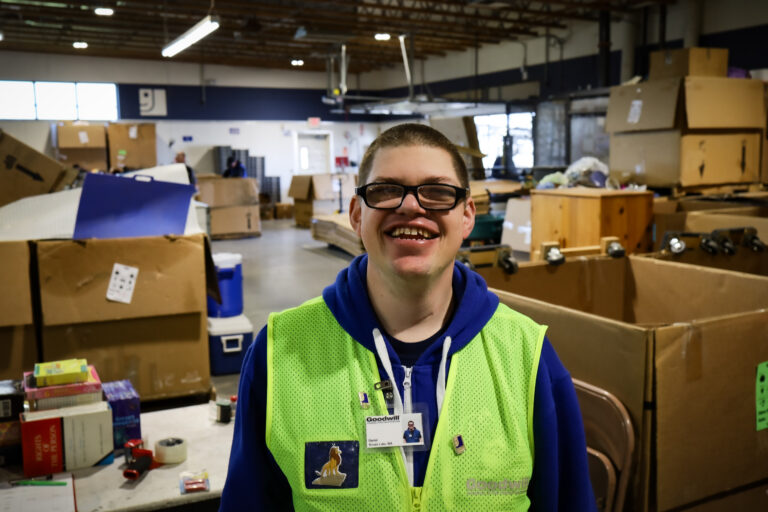 A man wearing glasses and a bright yellow visibility vest is standing in a warehouse and smiling at the camera.