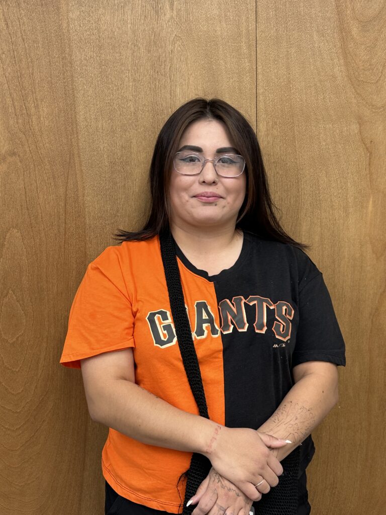 A woman wearing glasses and an orange and black "Giants" sports jersey is standing in front of a wooden wall and smiling at the camera.