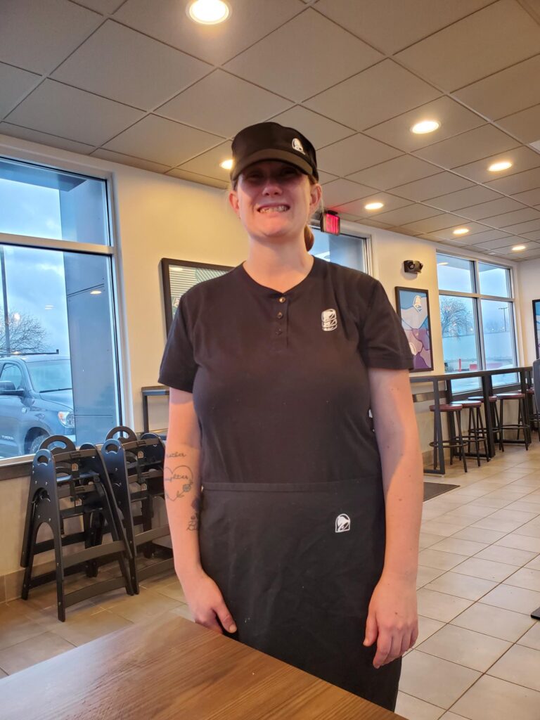 A woman in an all black uniform is standing in a restaurant dining area and smiling at the camera.