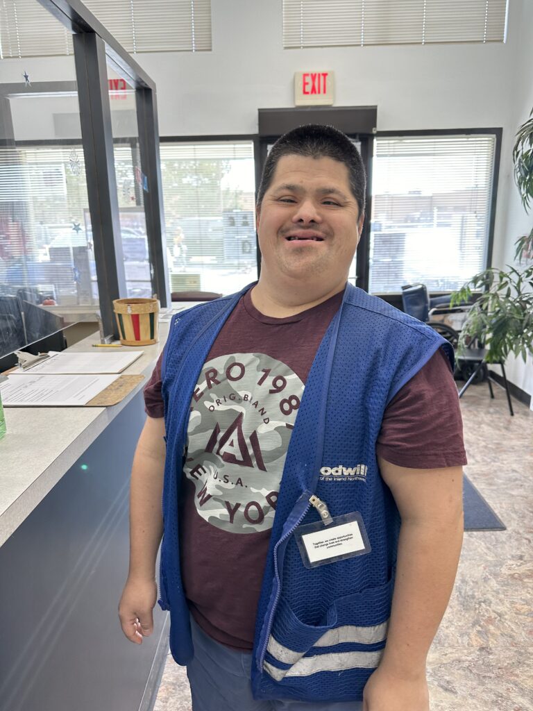 A man wearing a blue vest is standing in a lobby area and smiling at the camera.