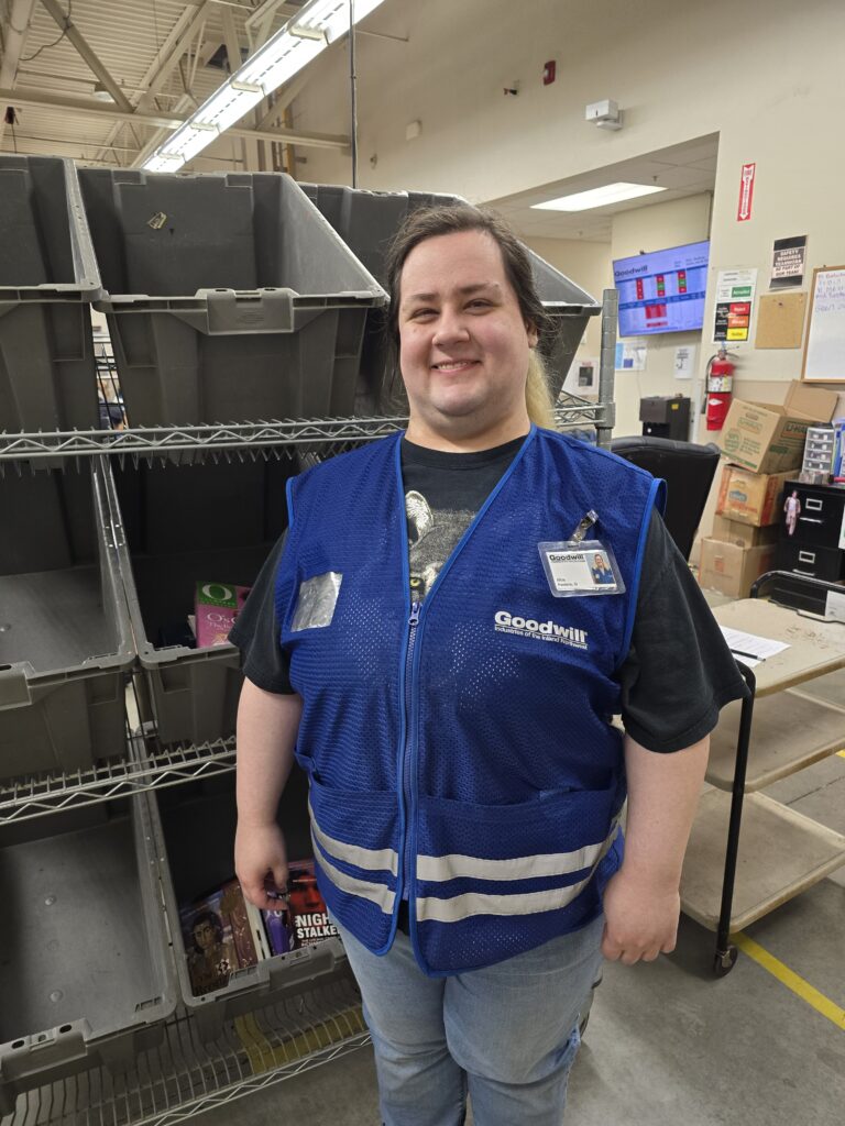 A woman wearing a blue vest is standing in a production area and smiling.