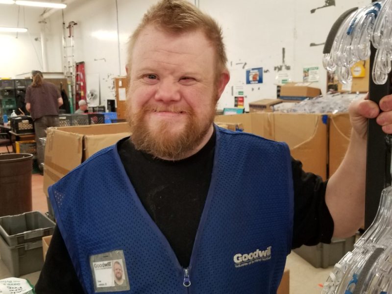 A man wearing a blue work vest with the word "Goodwill" on it, stands with his left hand gripping a textiles rack with empty hangers on it. He is smiling.