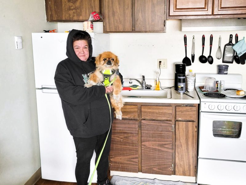 A woman holds a small dog and stands in a modest kitchen.